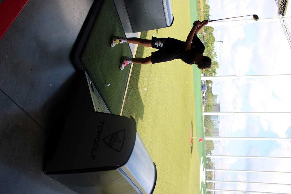 Group of smiling kids sit on couches at Topgolf holding golf clubs between turns.
